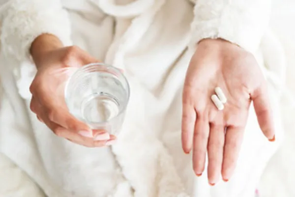 Женщина в одной руке держит стакан с водой, в другой - две капсулы